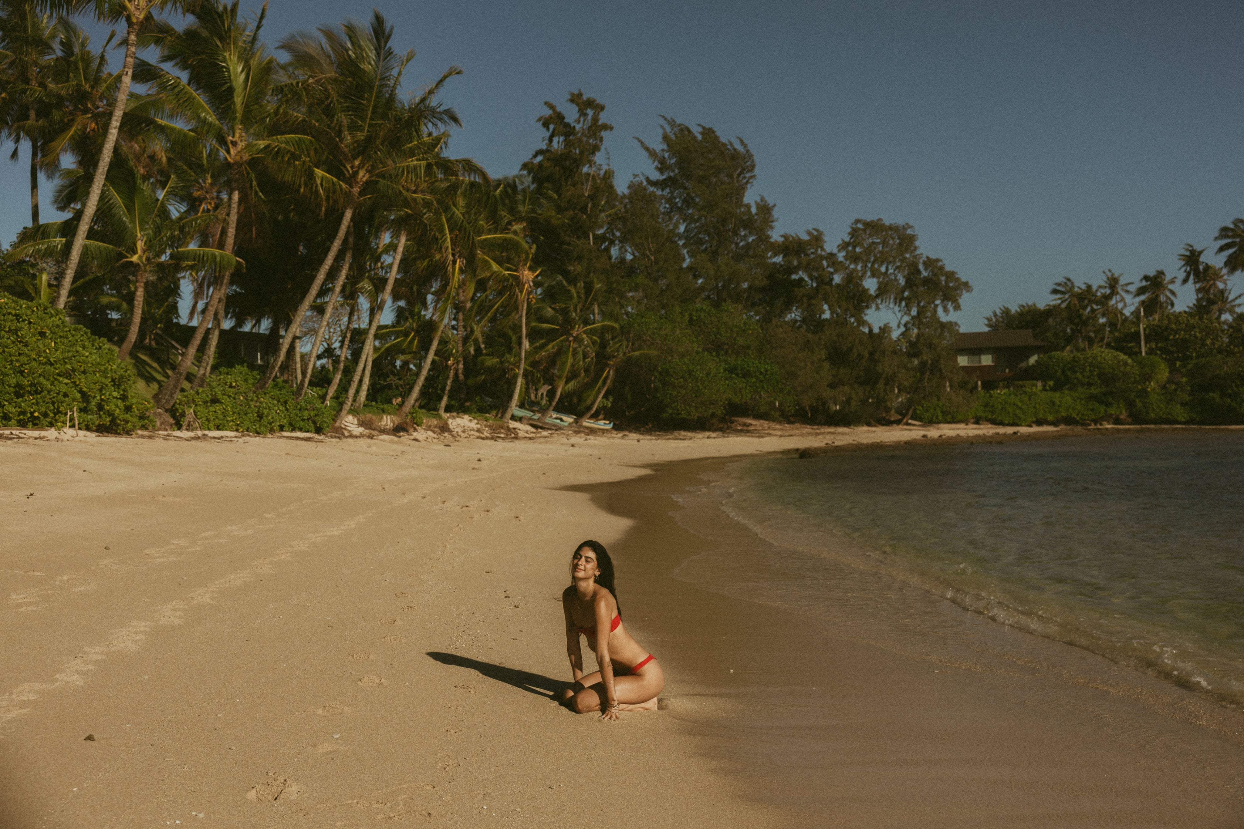Woman in a bikini sitting on a sandy beach with palm trees and water in the background